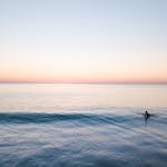 A lone surfer catches a wave at dawn, with a serene and pastel sky.